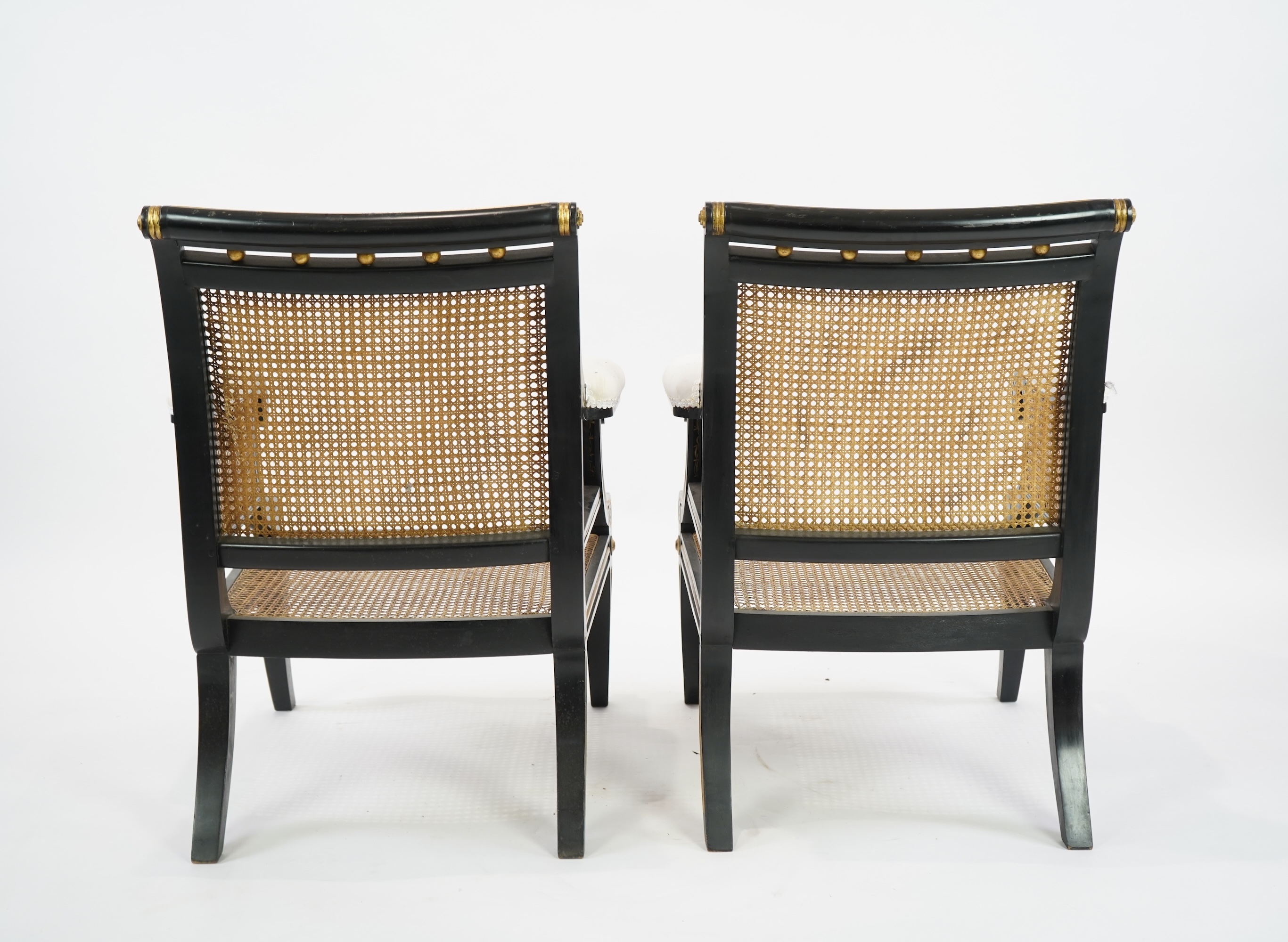 A pair of Regency style ebonised, parcel gilt caned bergere elbow chairs